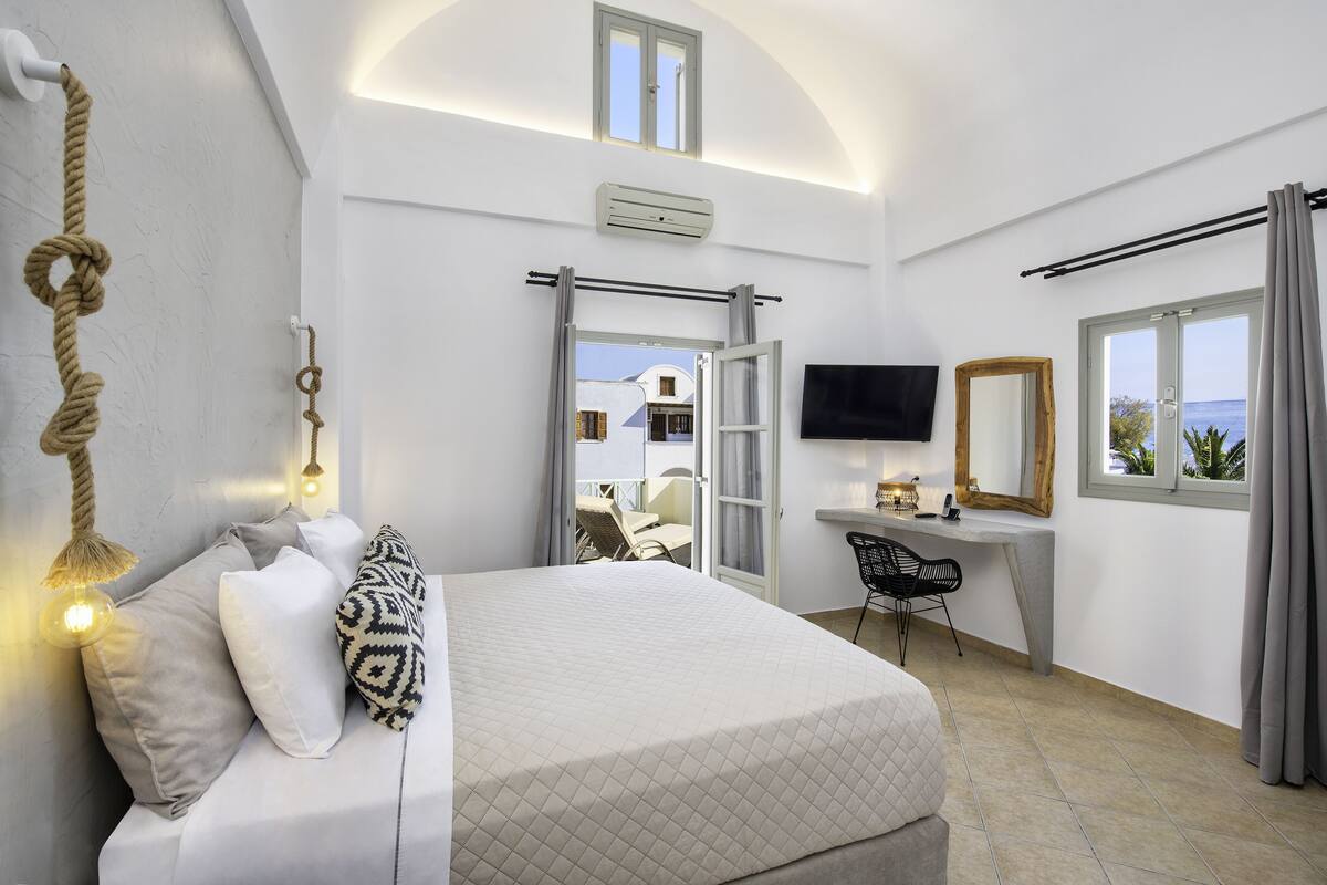 A bedroom with a bed, a desk, a chair, a TV, and a view of the sea.