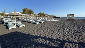 A beach with rows of white lounge chairs and thatched-roof structures. - Kalya Suites (Santorini)