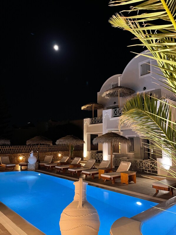A nighttime view of a resort pool area with lounge chairs and umbrellas, a large decorative vase, and a building with balconies. - Kalya Suites (Santorini)