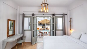 A hotel room with a bed, a desk with a chair, a mirror, and a view of an outdoor seating area through open doors. - Kalya Suites (Santorini)