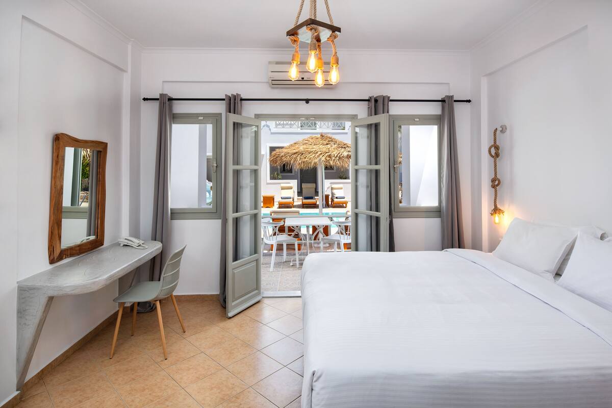 A hotel room with a bed, desk, chair, and a view of an outdoor dining area.