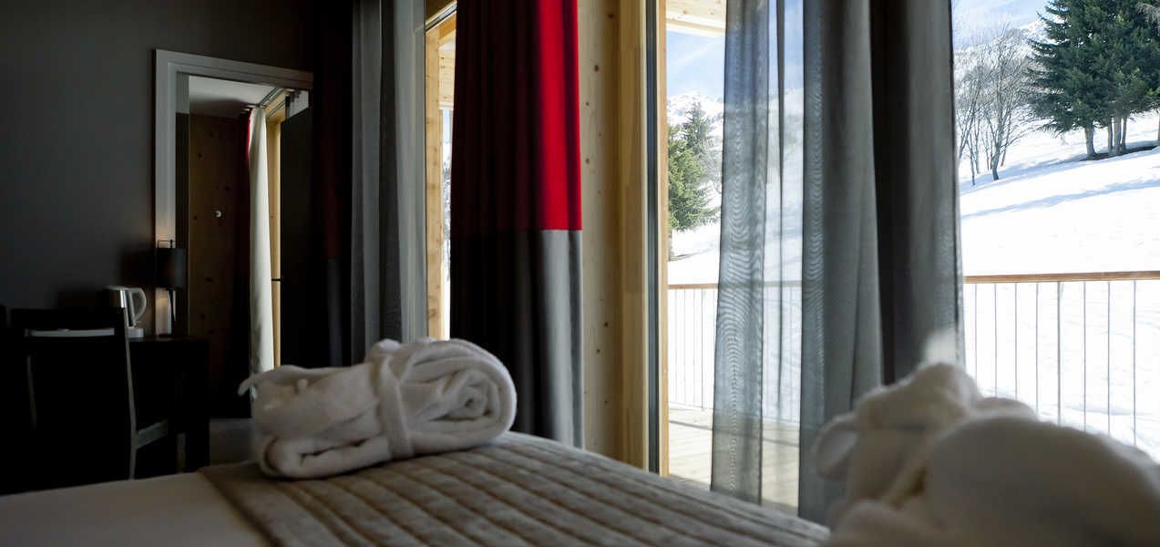 Une chambre d’hôtel avec un lit, une serviette soigneusement pliée, une fenêtre avec des rideaux et une vue sur un paysage enneigé.