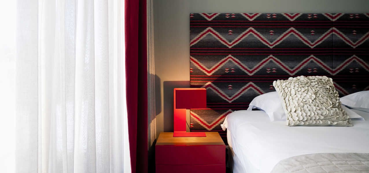 A hotel room with a bed, a nightstand, a red lamp, and a patterned headboard.