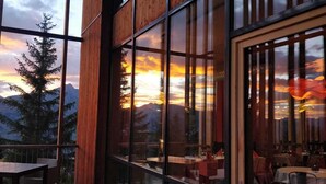 A restaurant with large windows offering a view of mountains and a sunset. - Aiguille Grive Chalets Hôtel (Bourg-Saint-Maurice)