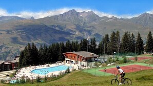 A mountain resort with a swimming pool, tennis courts, and a cyclist on a path. - Aiguille Grive Chalets Hôtel (Bourg-Saint-Maurice)