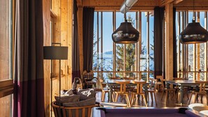 A wooden interior with a pool table, dining area, and large windows offering a view of snow-capped mountains. - Aiguille Grive Chalets Hôtel (Bourg-Saint-Maurice)