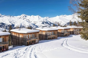 File d'habitacions de fusta amb teulades cobertes de neu, amb muntanyes nevades al fons.