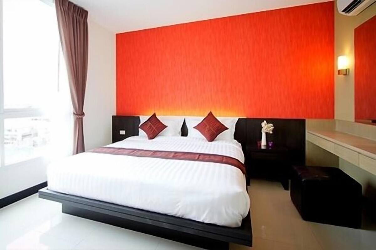 A modern hotel room with a large bed, a red accent wall, and a view of the city.