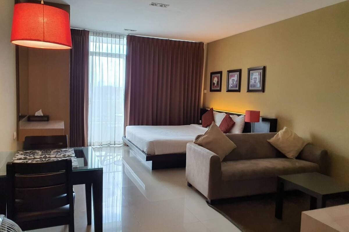 A hotel room with a bed, a sofa, a table, and a chair. There is a window with curtains, a red lamp, and framed pictures on the wall.