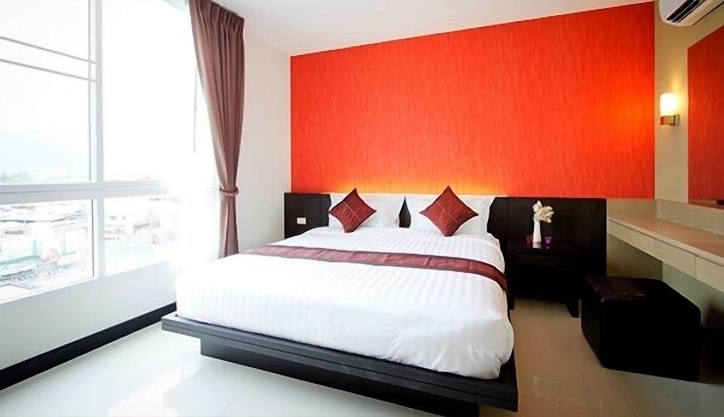 A modern hotel room with a large bed, a red accent wall, and a view of the city.