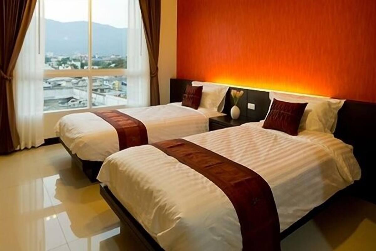 A hotel room with two beds, a large window with a view of a cityscape, and a red accent wall.