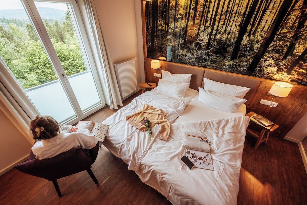 A bedroom with a bed, a nightstand, a lamp, a window with a view of a forest, and a chair with a person sitting.