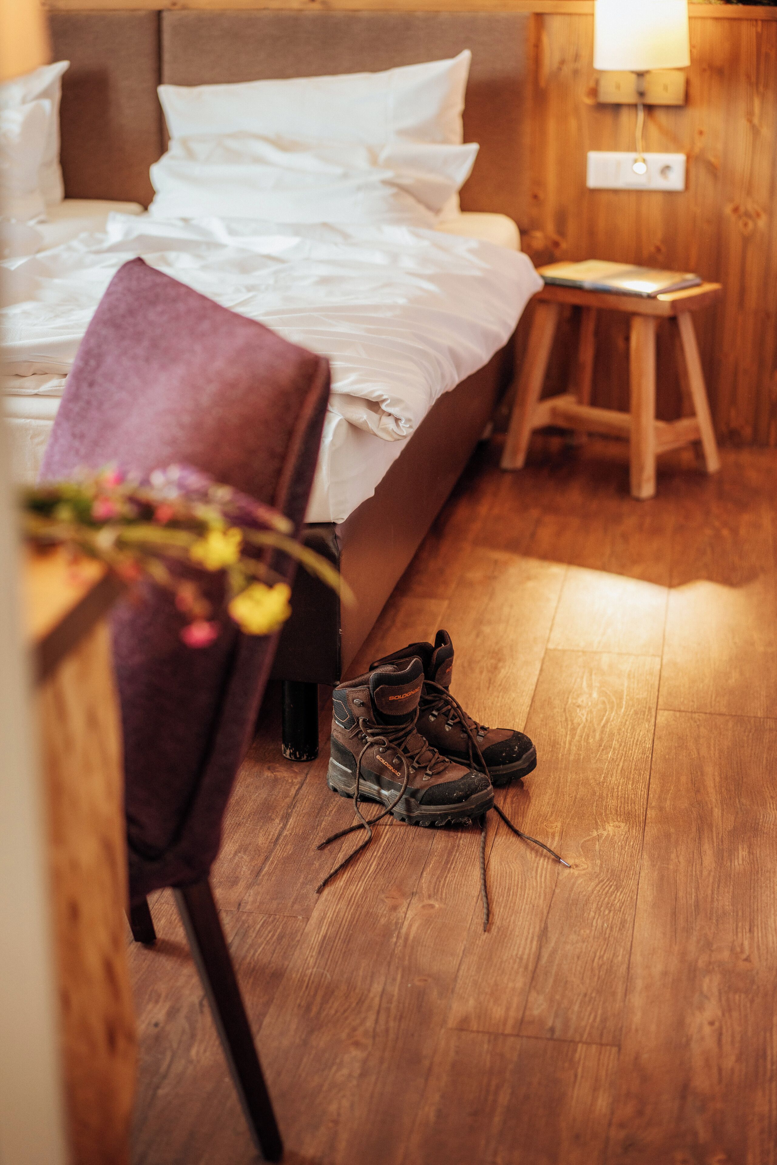 A bedroom with a wooden floor, a bed with white linens, a purple chair, and a pair of boots.