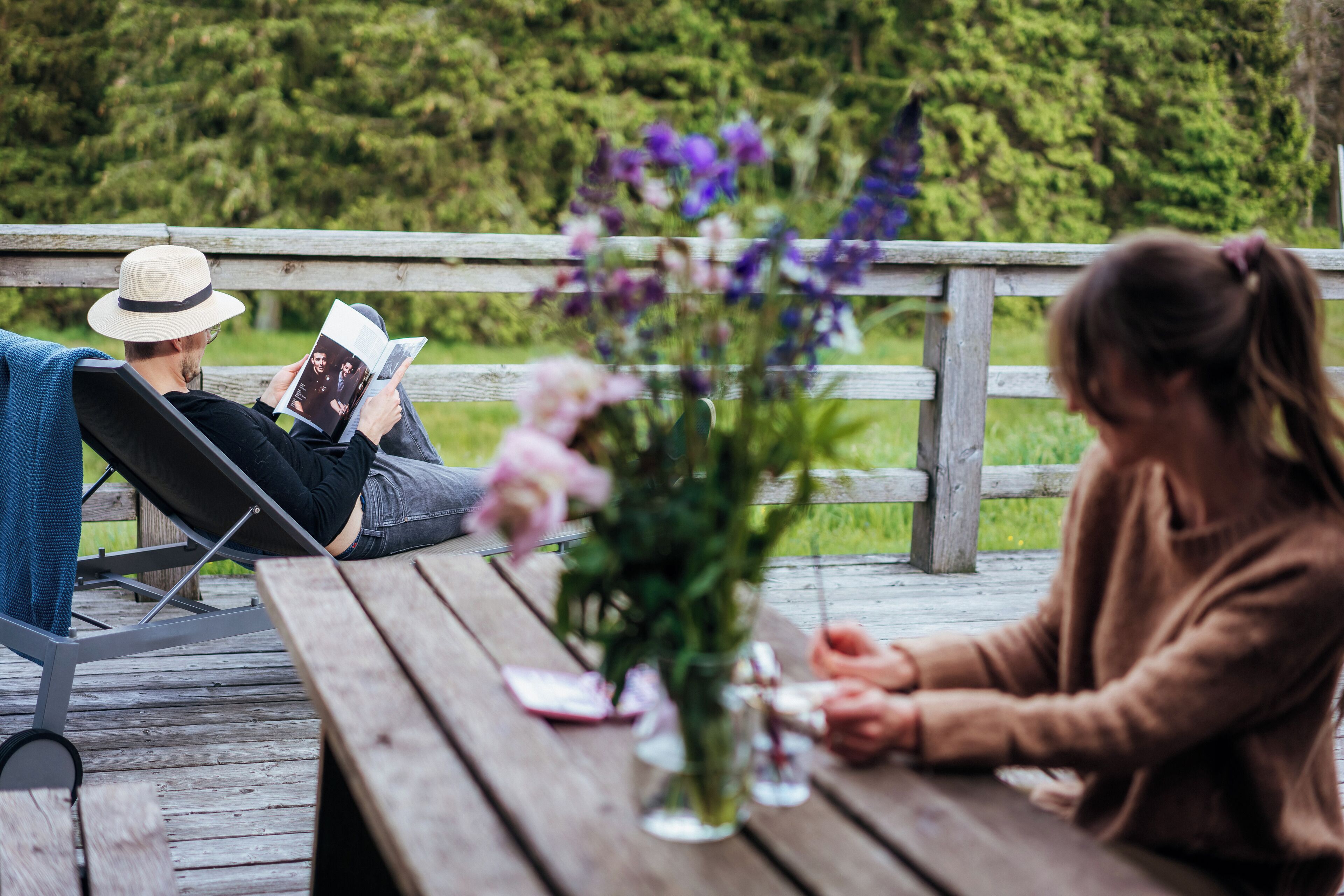 Eine Holzterrasse mit Picknicktisch, einer Blumenvase und Personen, die sitzen und lesen.