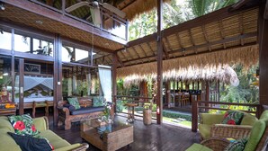 A spacious indoor area with wicker furniture, a glass coffee table, and a thatched ceiling. - Villa Kupu-Kupu (Ubud)