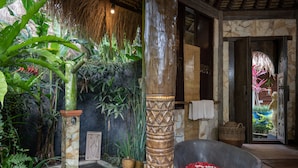 Un jardín tropical con un pilar de piedra, una fuente de piedra con flores rojas y una estructura cubierta de paja.