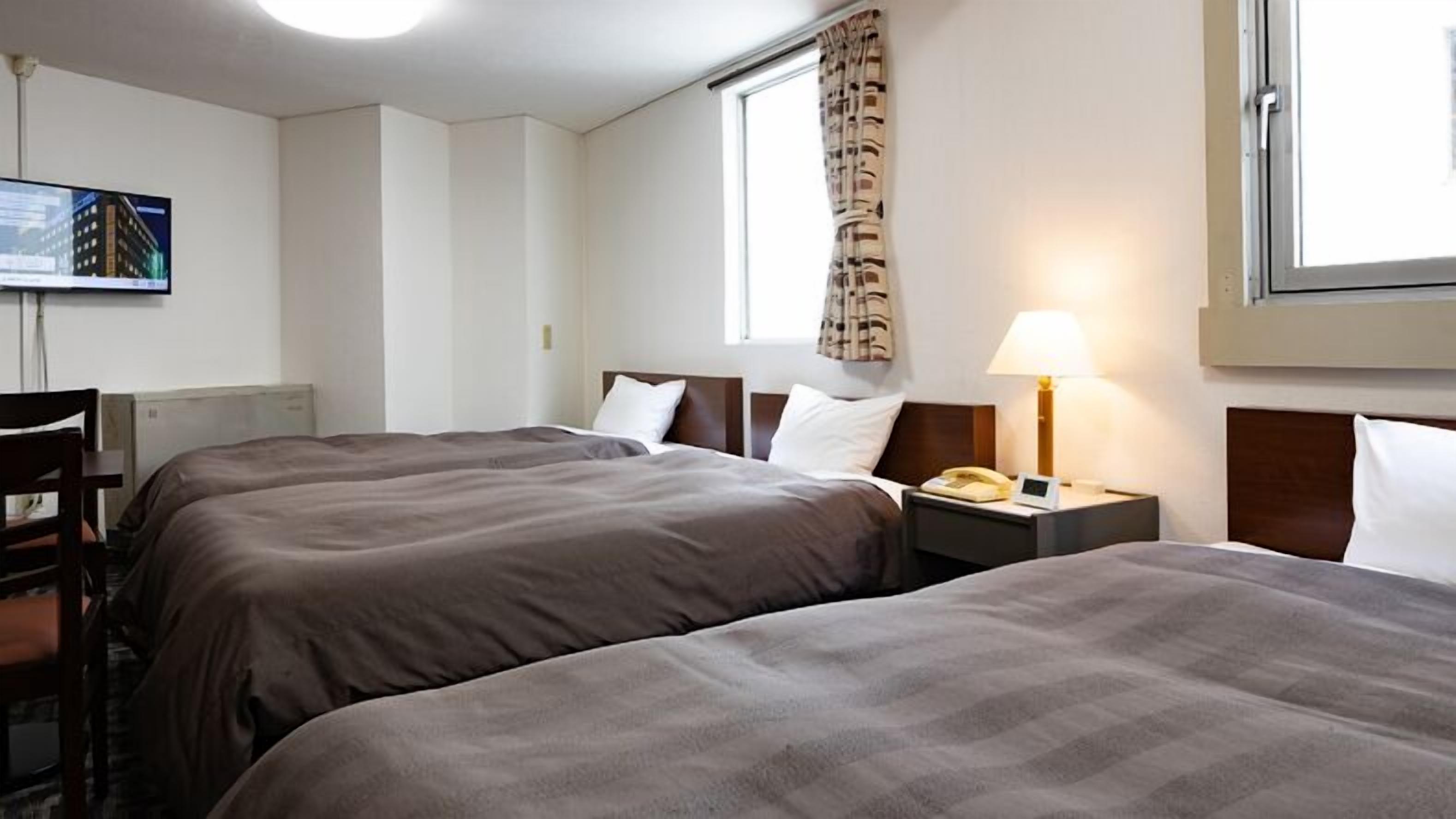 Ett hotellrum med två sängar, ett skrivbord, en stol, en lampa, en telefon och ett fönster med gardiner.