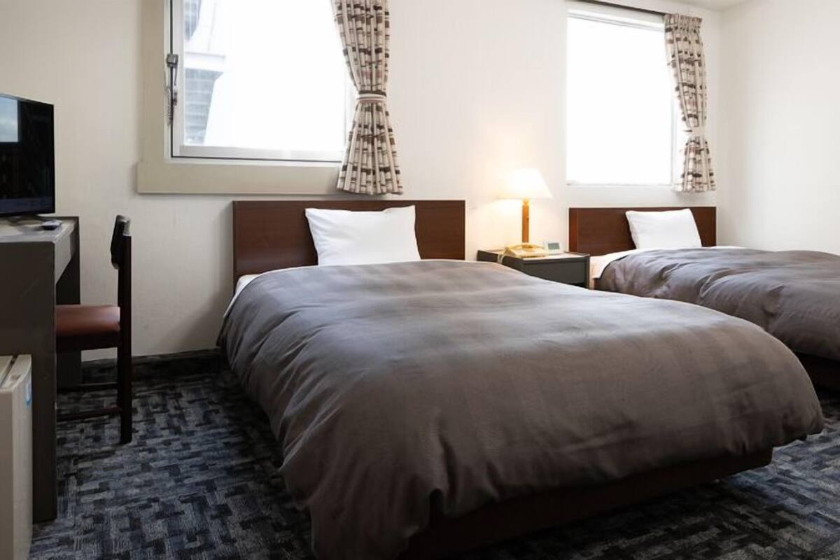 Ein Hotelzimmer mit zwei Betten, einem Schreibtisch mit Computer, einem Stuhl und einem Fenster mit Vorhängen.