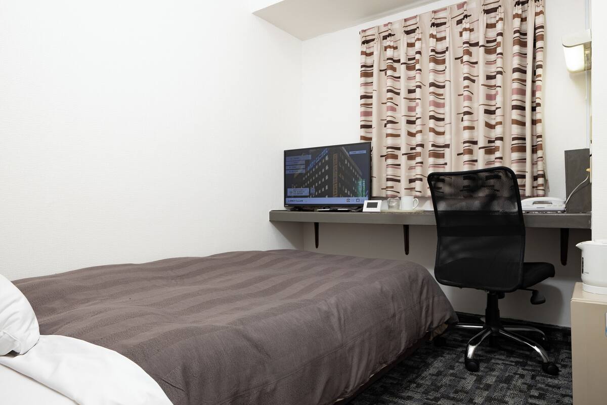 Ein Hotelzimmer mit Bett, Schreibtisch mit Computer, Stuhl und einem Fenster mit Vorhängen.