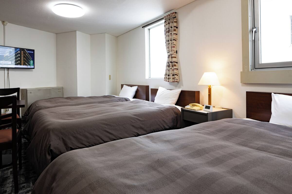 Ein Hotelzimmer mit zwei Betten, einem Schreibtisch mit Lampe, einem an der Wand montierten Fernseher und einem Fenster mit Vorhängen.
