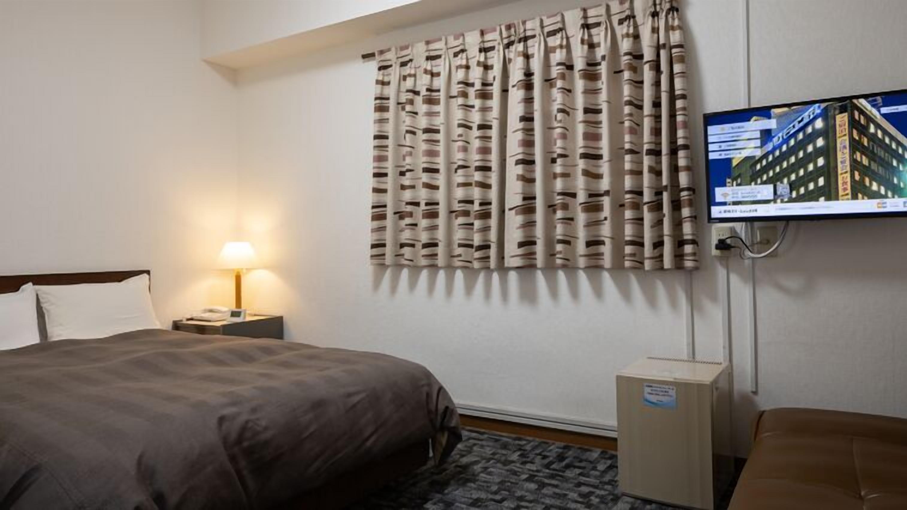 Ett hotellrum med en säng, ett nattduksbord med en lampa, en tv som är monterad på väggen och ett fönster med gardiner.