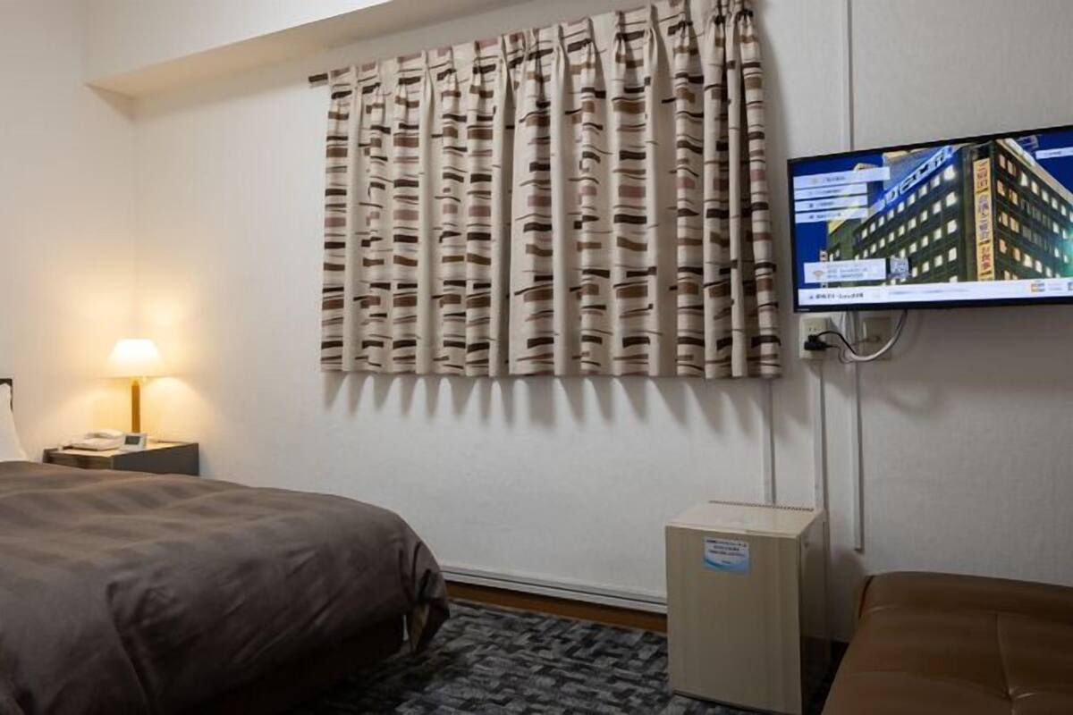 Ein Hotelzimmer mit Bett, Nachttisch mit Lampe, an der Wand montiertem Fernseher und einem Fenster mit Vorhängen.