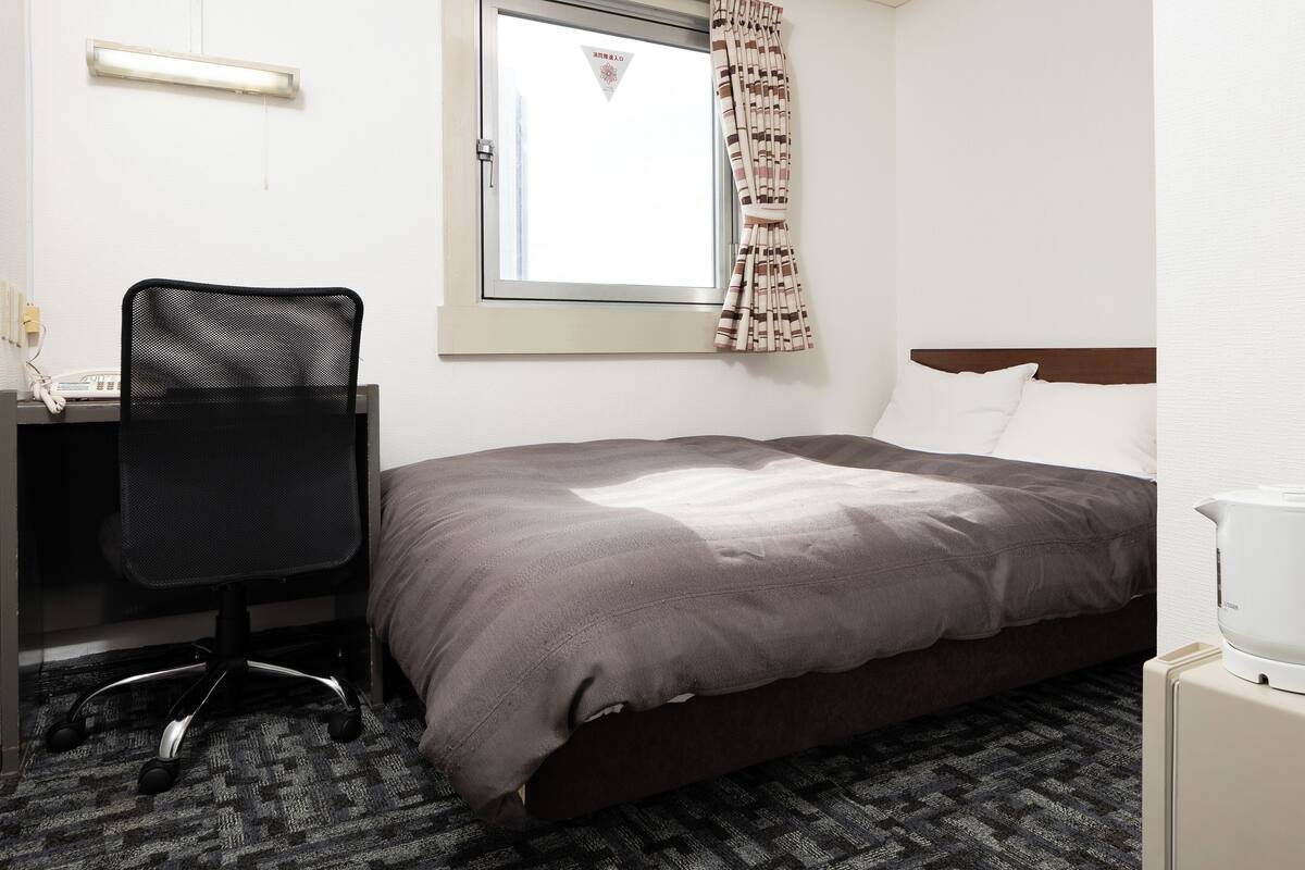 Ein Hotelzimmer mit Bett, Schreibtisch mit Stuhl, Fenster mit Vorhängen und Nachttisch mit Lampe.
