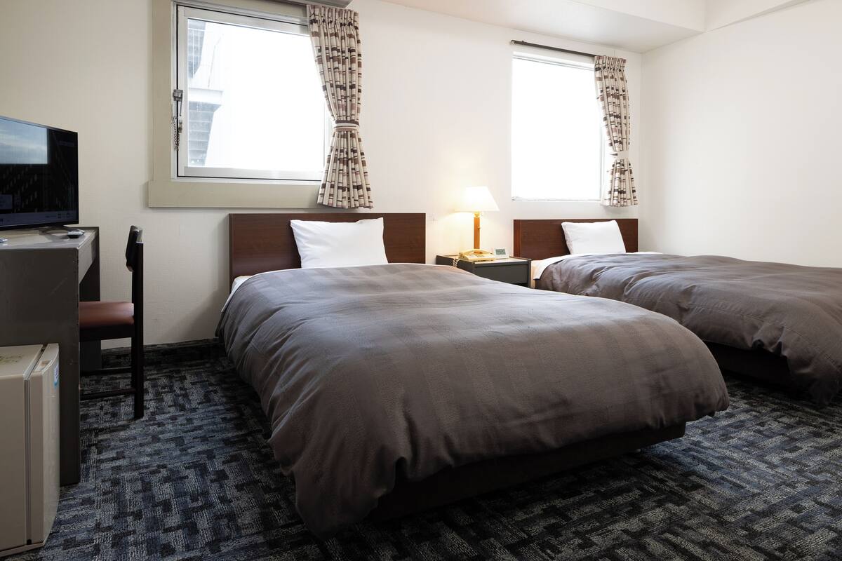 Ein Hotelzimmer mit zwei Betten, einem Schreibtisch mit Fernseher und einem Fenster mit Vorhängen.