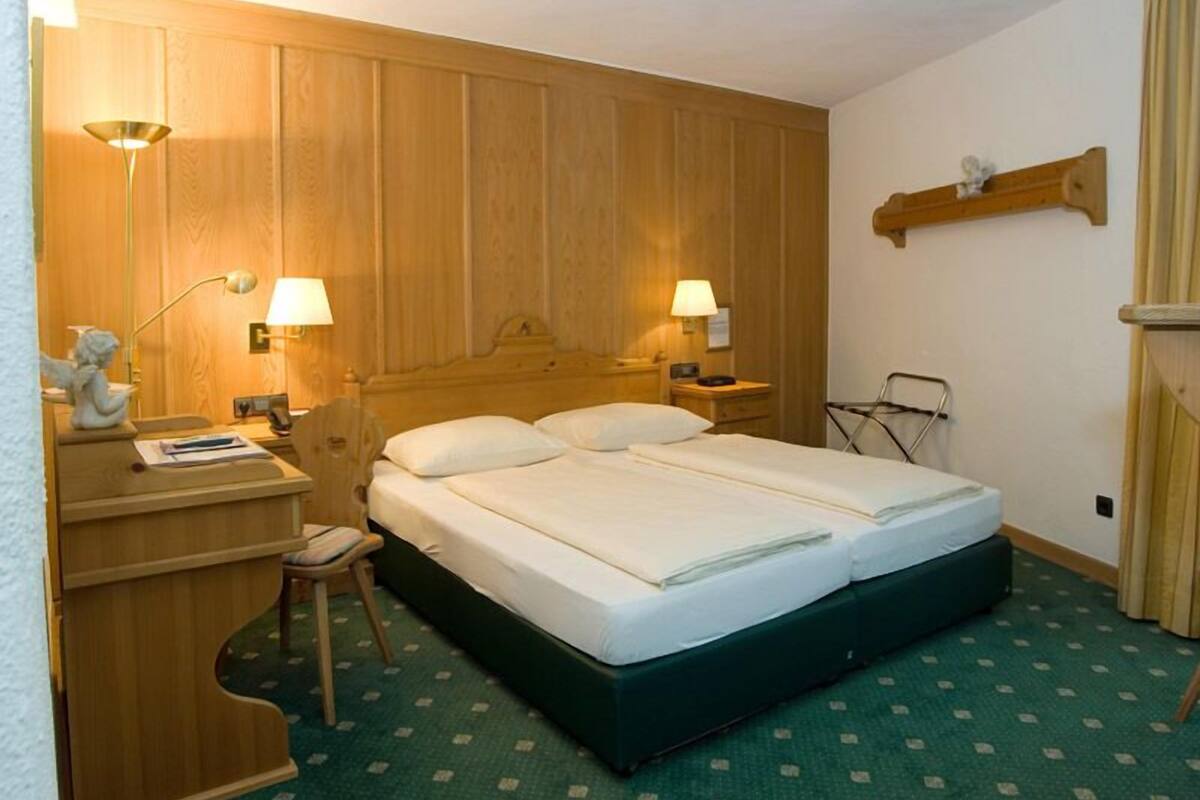 A hotel room with a double bed, wooden headboard, two bedside lamps, a desk with a lamp, a chair, and a small shelf.