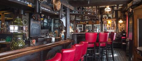 A bar with wooden beams, red bar stools, and various bottles and glasses on shelves.