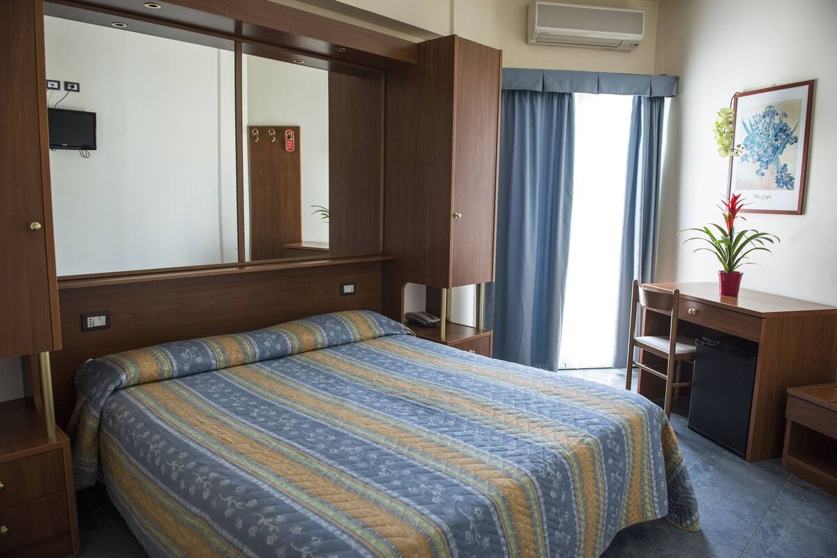 A hotel room with a large bed, a desk, a chair, a small fridge, a potted plant, and a window with curtains.