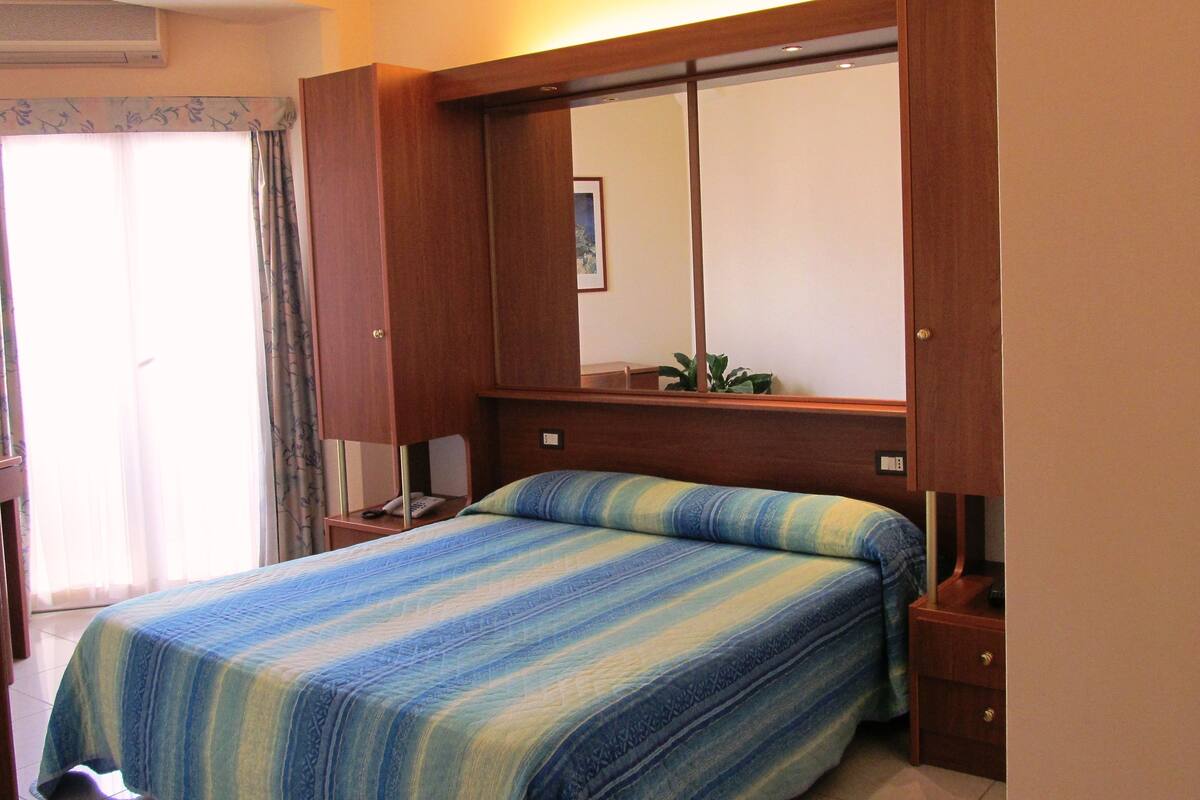 A bedroom with a bed, a nightstand, a mirror, and a window with curtains.
