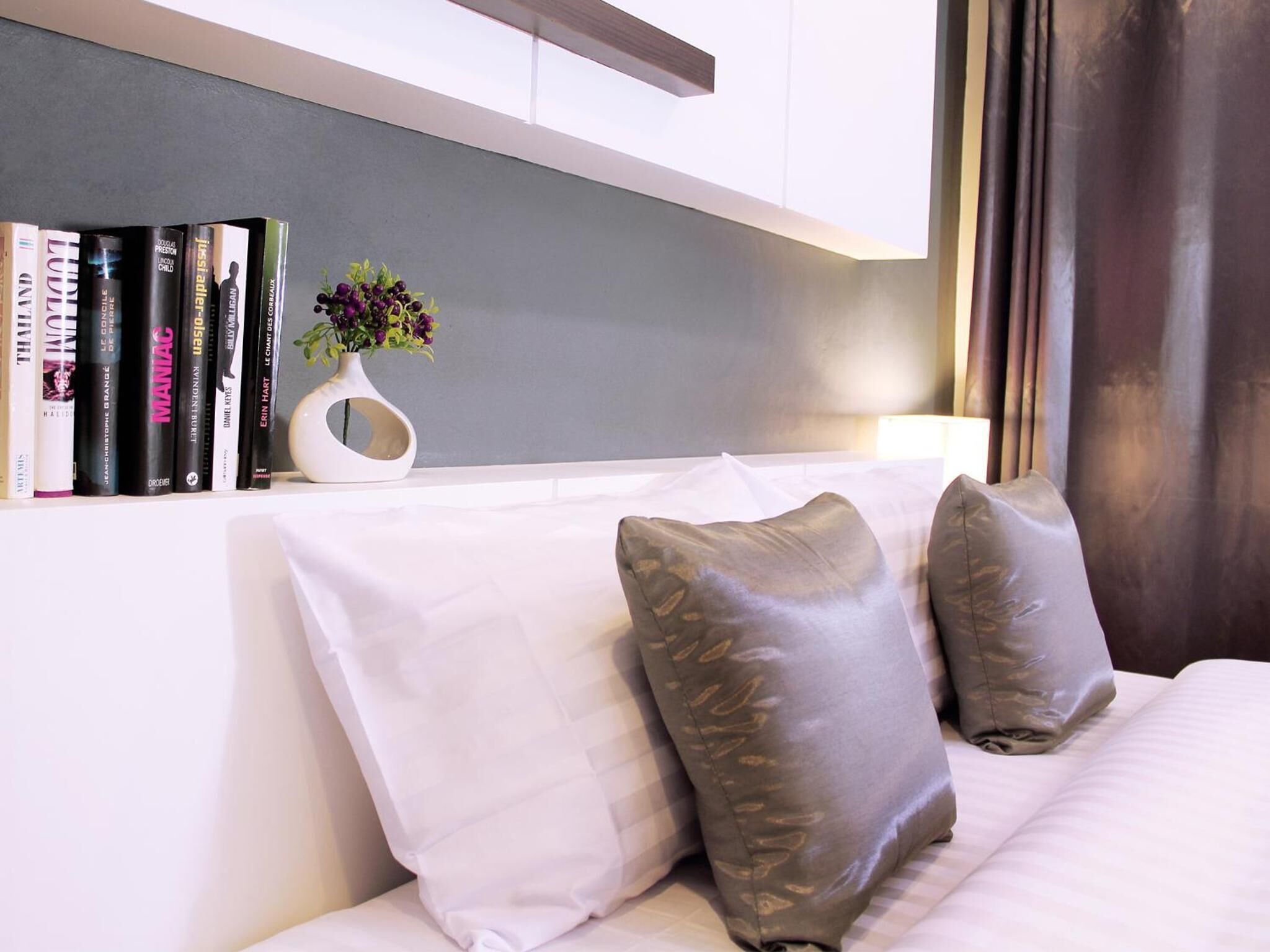 Une chambre d’hôtel moderne, avec une bibliothèque bien rangée, un vase blanc orné de fleurs et un lit avec des oreillers gris.