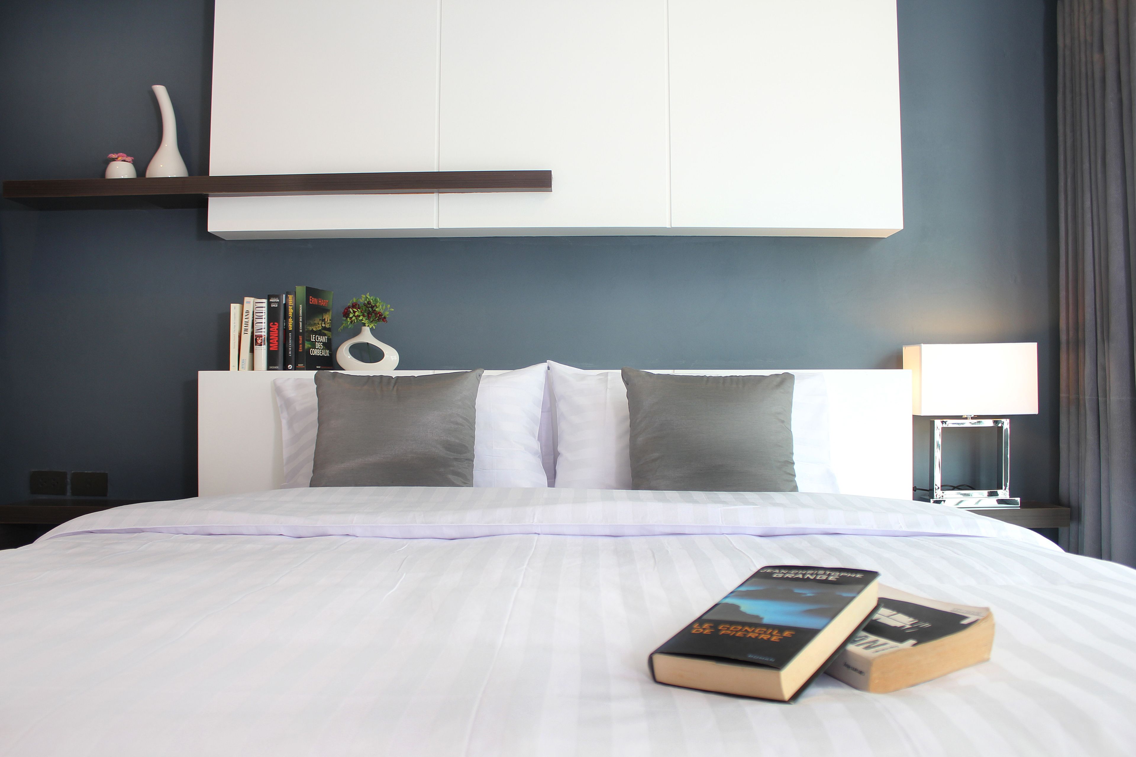 Una cama bien hecha con una colcha blanca y almohadas, una mesita de noche con una lámpara, y una estantería con libros y un florero.