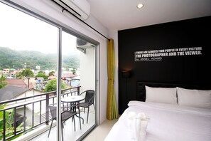 Une chambre d’hôtel avec un balcon, un lit avec du linge de lit blanc, une tête de lit noire et une citation affichée sur le mur.