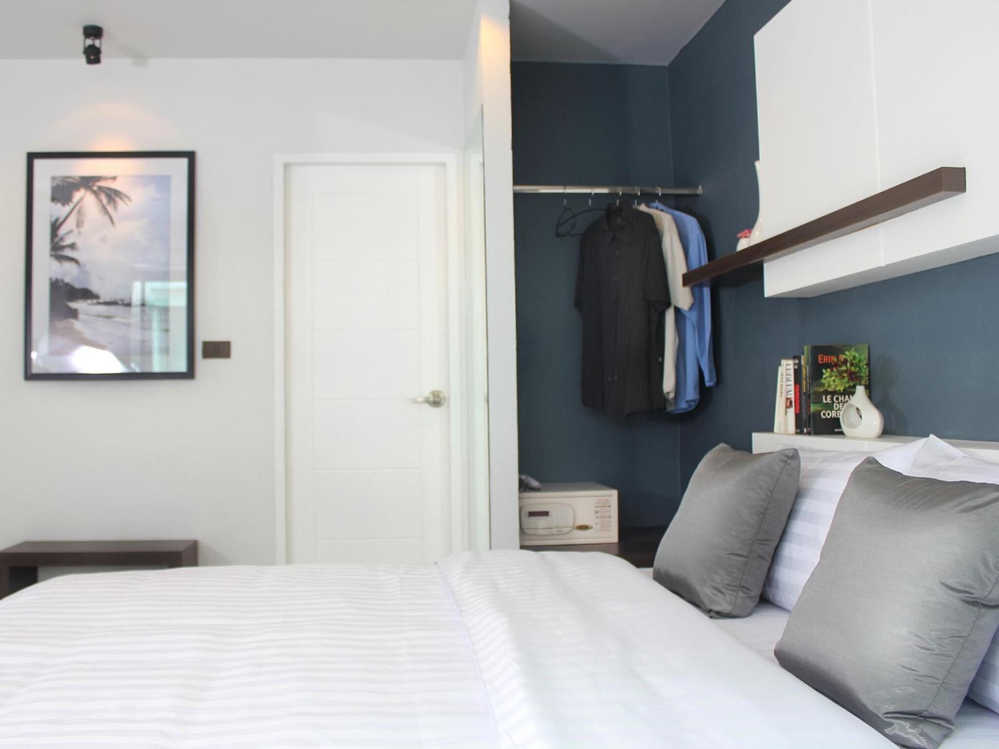Un dormitorio con una cama, almohadas, una mesita de noche, un perchero con ropa, un cuadro en la pared y una mesita auxiliar con un libro y un jarrón.