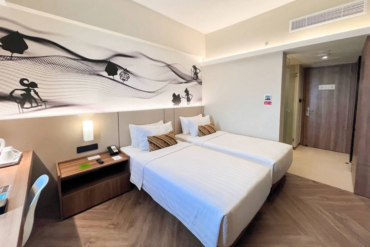 A hotel room with two beds, a desk, a chair, a wall-mounted artwork, and a door.