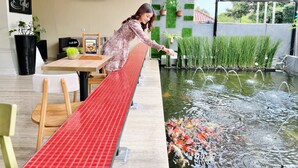 Una mujer con un vestido estampado se apoya en un banco de tejas rojas junto a un estanque de carpas, con un cartel de 'Zuri Lounge' al fondo.