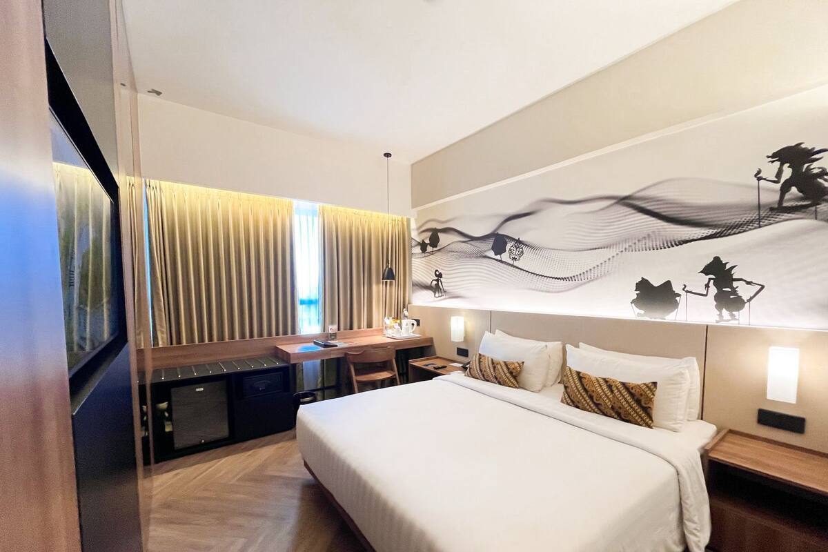 A modern hotel room with a large bed, a desk, a chair, and a small kitchenette.
