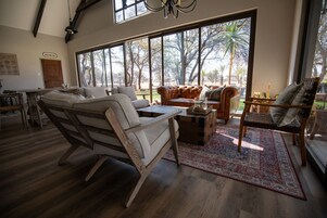 Amplia sala de estar con ventanas grandes, suelo de madera y diversas opciones de asientos, incluyendo un sofá y sillones.