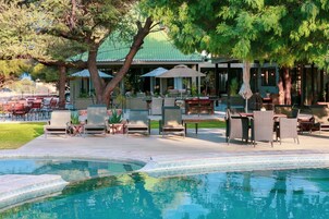 Un área de piscina con sillones de descanso y sombrillas, una zona de estar cubierta y un edificio con techo verde al fondo.
