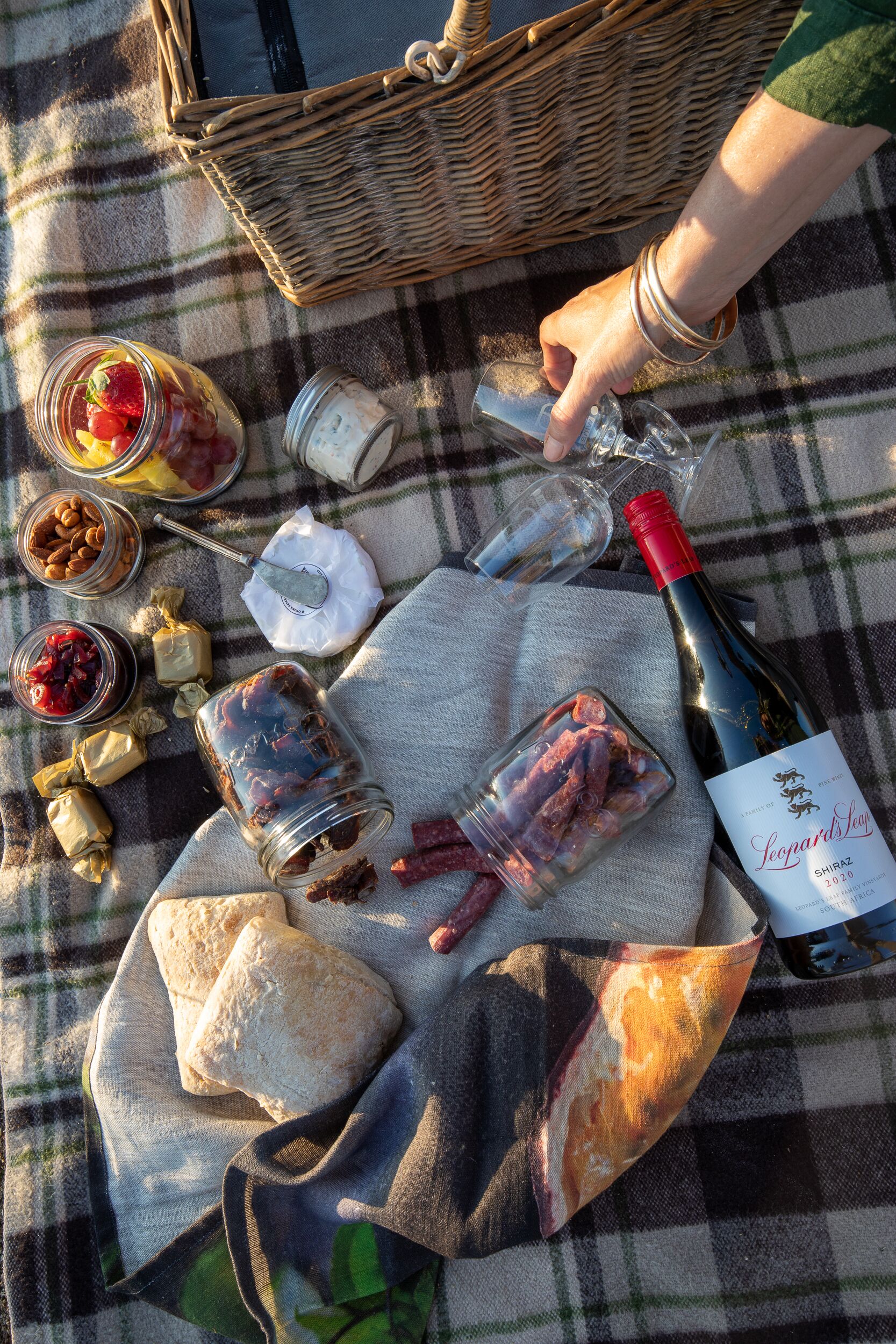 Un montaje para picnic con una botella de vino, frascos de comida y una cesta sobre un mantel a cuadros.