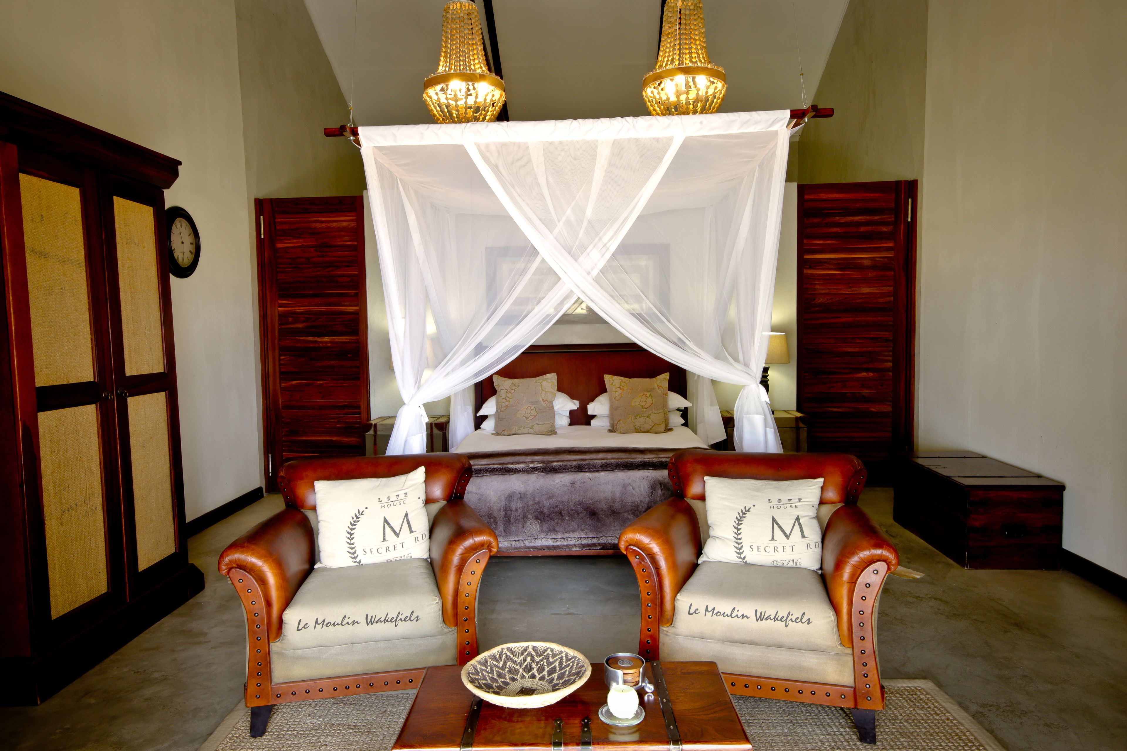 Un dormitorio con cama con dosel, dos sillones de cuero, una mesita de noche con una vela y un cuenco decorativo.
