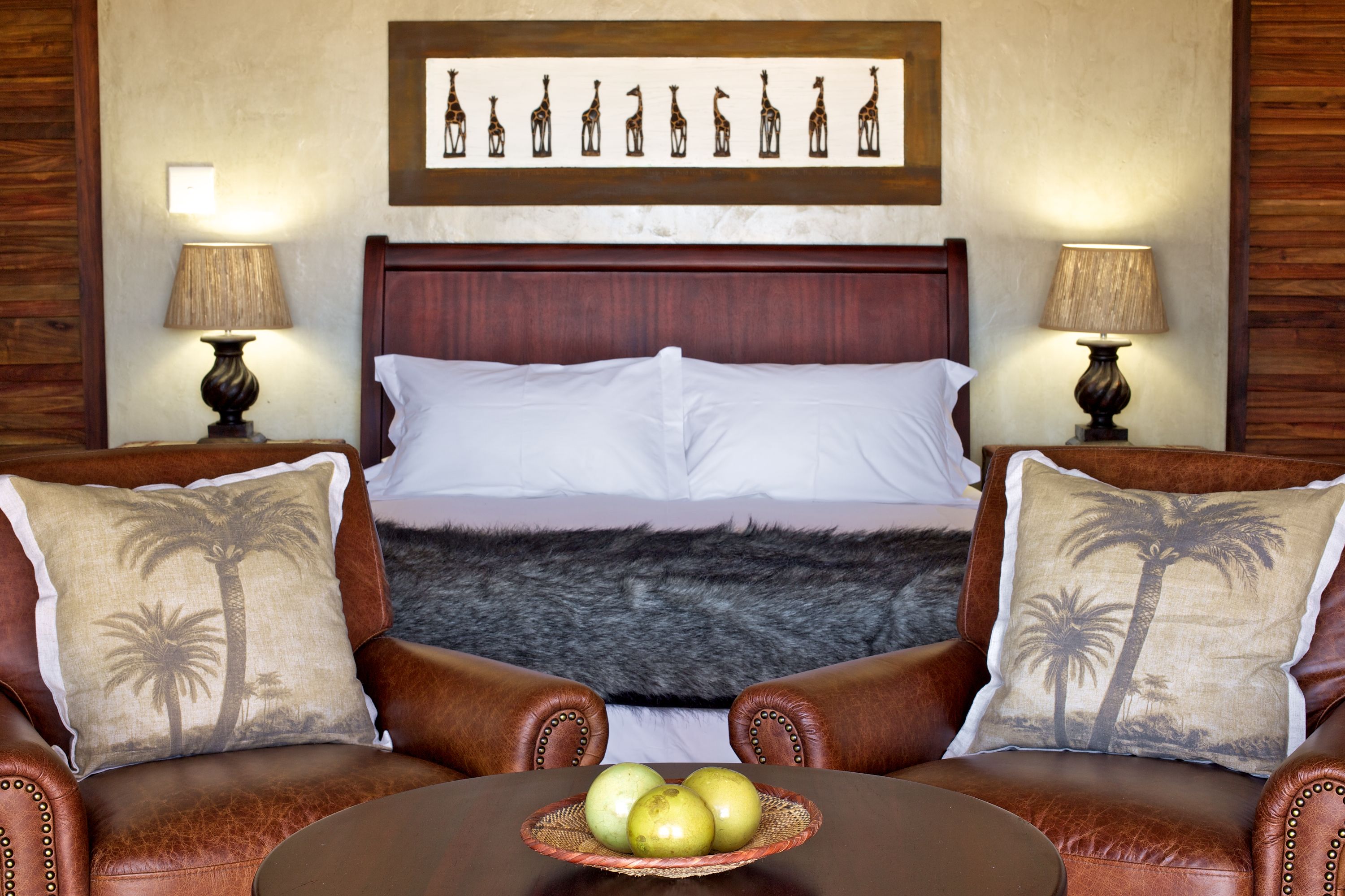 A hotel room with a large bed, two leather armchairs, a round table with apples, and a wall with a giraffe mural.