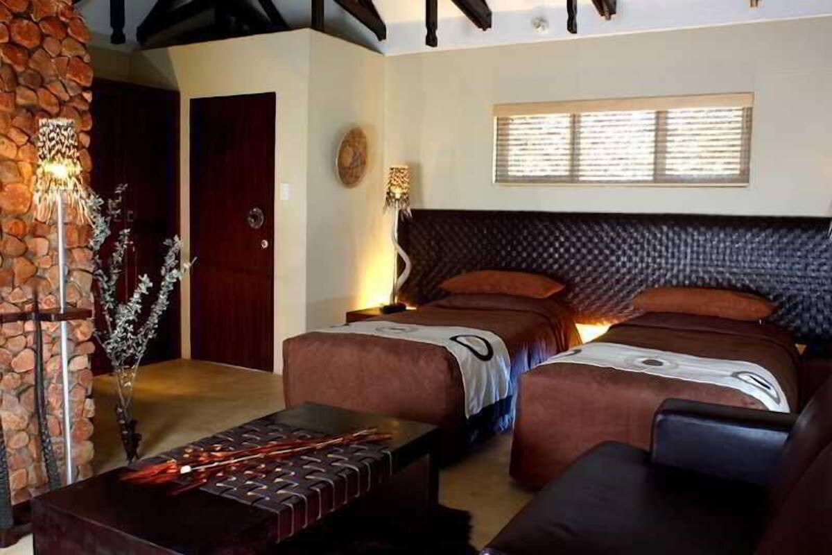 Une chambre avec deux lits, un mur en pierre, un plafond en bois et une fenêtre avec des stores.