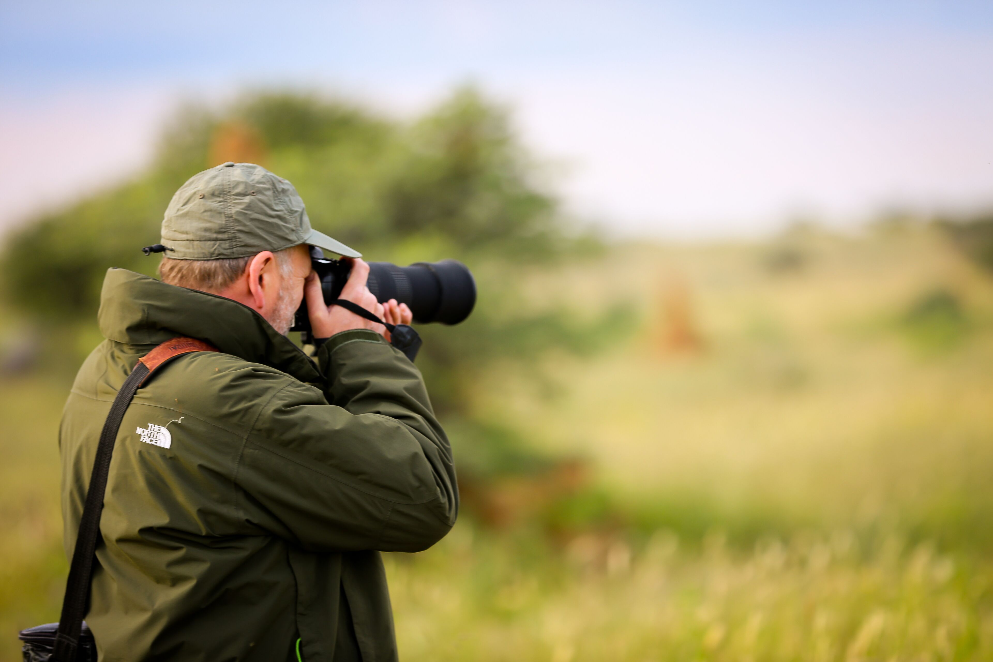 A person in a green jacket and cap taking a photo with a large camera in an outdoor setting.