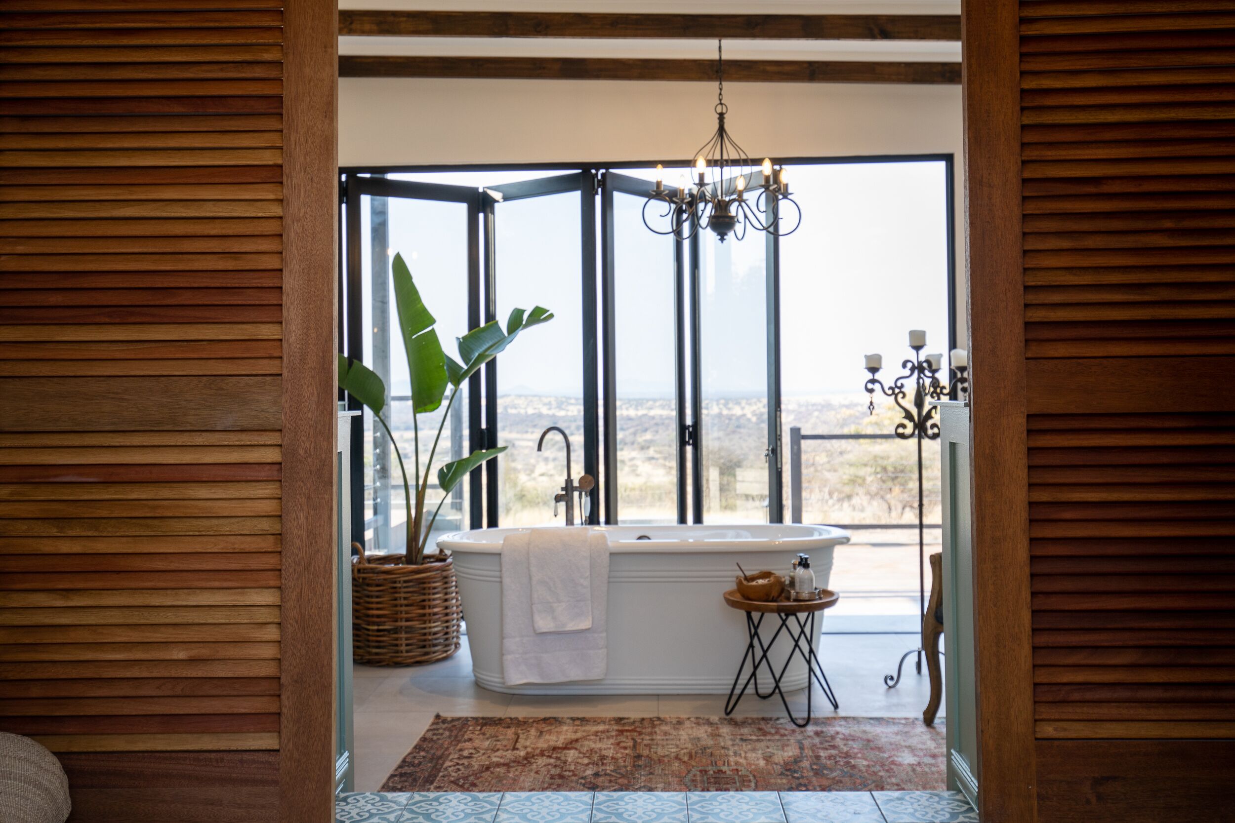 A bathroom with a freestanding bathtub, a large window, a chandelier, and a potted plant.