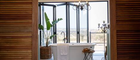A bathroom with a freestanding bathtub, a large window, a chandelier, and a potted plant.