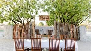 A wooden dining table set with white tablecloths and chairs, surrounded by a rustic stone wall and trees. - Ongula Village Homestead Lodge (Ondangwa)