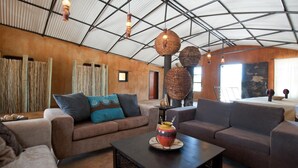A spacious room with a high ceiling, featuring a large window, a sofa set, and a coffee table with a decorative vase. - Ongula Village Homestead Lodge (Ondangwa)